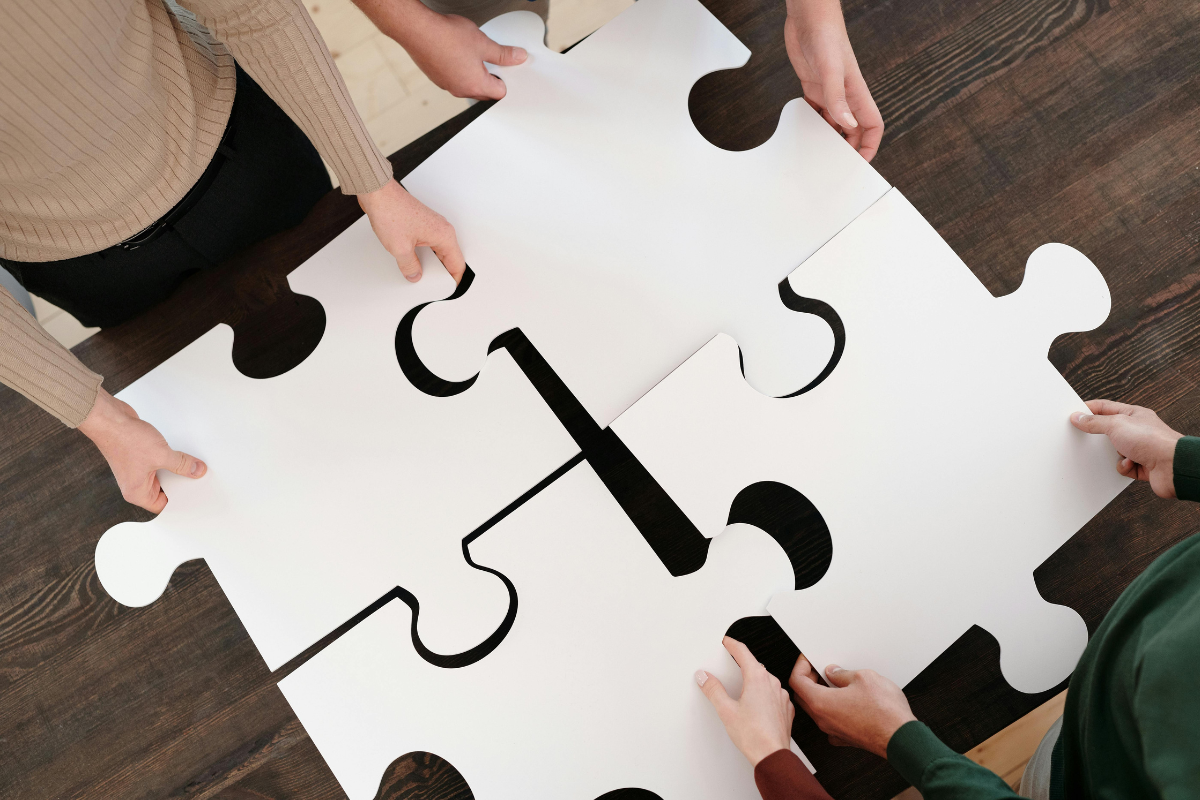 Four people are connecting oversized puzzle pieces on a wooden table, symbolising teamwork and collaboration.